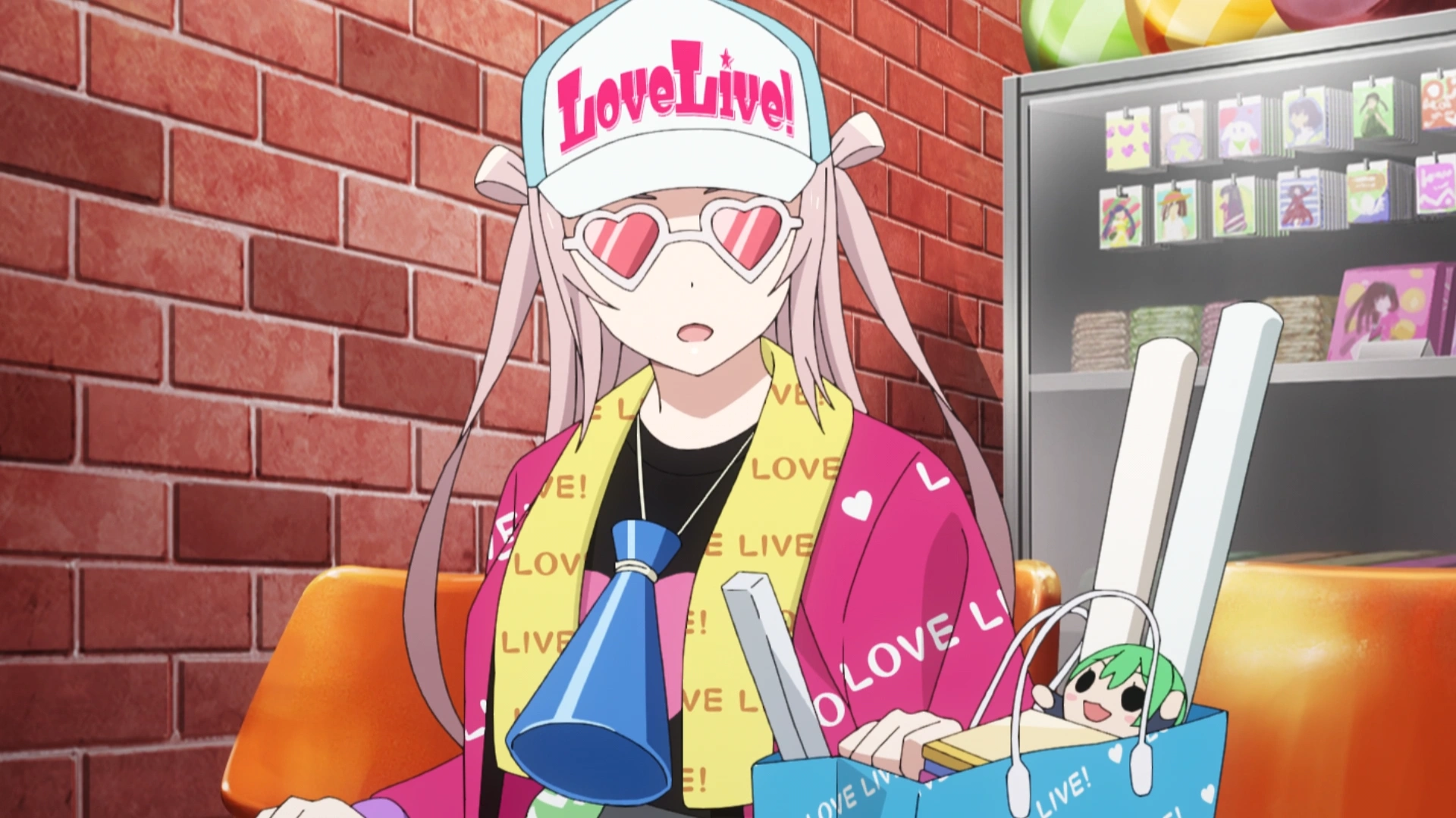 Lanzhu Zhong with heart glasses, and a black shirt with a heart on it. She has a cap on that says "LoveLive!" and various other items and clothing that says "LOVE LIVE." Various other goods can be seen on or around her. She is covered in merch.